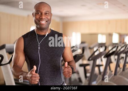 Ritratto di un uomo nero sorridente nel fitness club. Ha i pollici in su in atteggiamento positivo. Concetto di sport e vita sana. Foto Stock