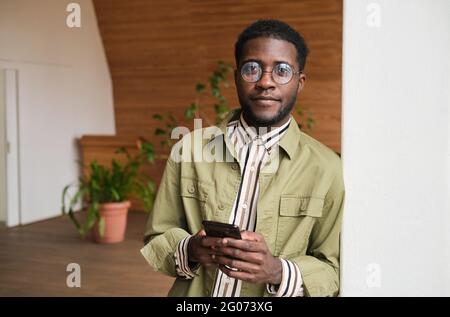 Ritratto di un giovane uomo d'affari africano che guarda la fotocamera e usa il telefono cellulare mentre si trova in ufficio Foto Stock