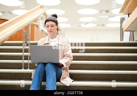 Giovane donna con un computer portatile in ginocchio che studia mentre si siede sulle scale Foto Stock