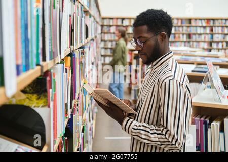 Studente africano in piedi davanti agli scaffali e leggendo un libro nella biblioteca Foto Stock