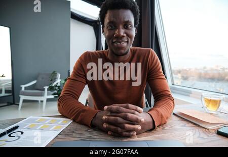 Ritratto di un giovane africano sorridente in camera mentre si siede sul posto di lavoro in ufficio Foto Stock