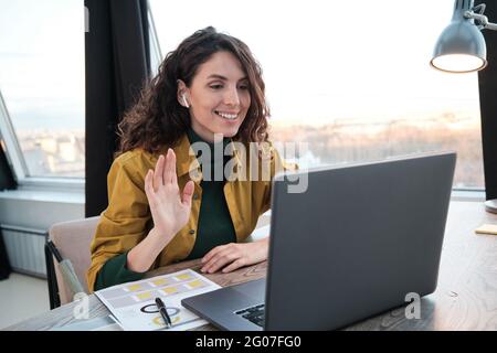 Giovane donna d'affari sorridente e sventolante mentre parla online con il suo collega sul portatile Foto Stock