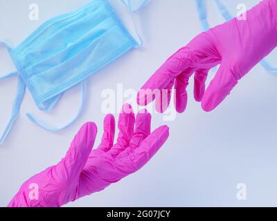 Due mani in guanti rosa in lattice che si allungano l'una verso l'altra. Maschera medica protettiva blu su sfondo bianco. Concetto di protezione antivirale. Foto Stock