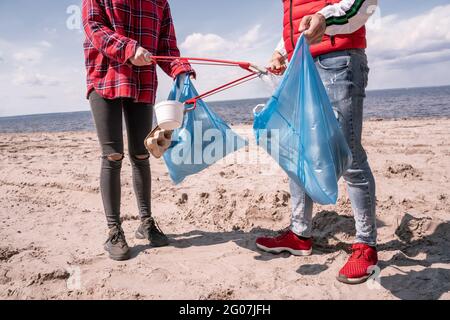 vista tagliata di coppia con sacchi di spazzatura e grabbers che raccolgono immondizia sulla sabbia Foto Stock