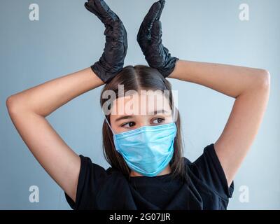Femmina adolescente ragazza in una maschera medica di protezione blu mostra corona tenendo le mani in nero guanti di lattice sopra la testa. Concetto di coronavirus Foto Stock