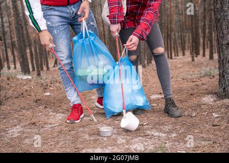 vista ritagliata di coppia con sacchi di spazzatura raccogliendo rifiuti con attrezzi di afferramento in foresta Foto Stock