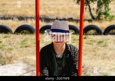 Defocalizzare la giovane donna sorridente in camicia a scacchi e cappello che oscillano sull'altalena sul parco giochi. Hippers e hippie ritratto. Zona di campagna. swi rosso brillante Foto Stock