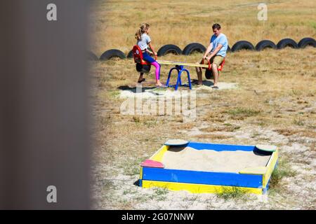 Defocus ragazza oscillare sul campo da gioco con il giovane uomo, ragazzo, suo fratello maggiore. Campagna. Swing e sandbox blu e rosso brillante. Estate in famiglia Foto Stock