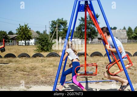 Defocalizzare la bambina che oscilla sul campo da gioco con il giovane uomo, ragazzo, suo fratello maggiore. Zona di campagna. Altalena blu e rossa. Estate in famiglia Foto Stock