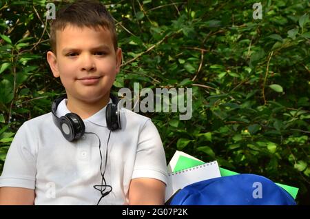 Un ragazzo di scuola in una camicia bianca accanto a una valigetta nel parco sta suonando musica con le cuffie. Studenti che imparano ad ascoltare i tutorial audio in un parco Foto Stock