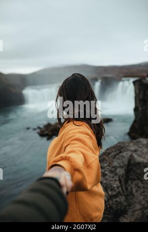 Vista laterale del giovane turista in occhiali con piercing holding mano di umano e guardando lontano sulla collina vicino alla cascata e fiume di montagna Foto Stock