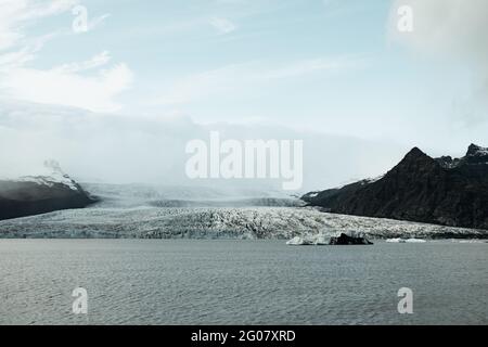 Vista pittoresca della superficie dell'acqua vicino a un enorme ghiacciaio incredibile tra colline rocciose e cielo blu Foto Stock