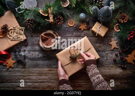 Vista dall'alto di una donna anonima che prepara un regalo di Natale accanto a una ciotola di cioccolato e decorazioni natalizie su un tavolo di legno Foto Stock