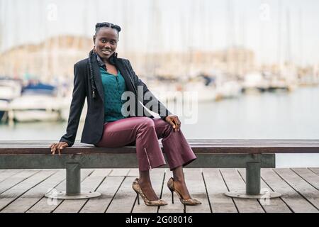 Elegante donna afro-americana sorridente trendy che passa il tempo sedendosi sopra panca bassa in legno nel parco in giorno luminoso Foto Stock
