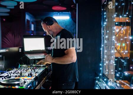 Vista laterale, uomo di mezza età in cuffia e nero Abbigliamento casual che mette a fuoco e gioca DJ controller professionale mentre si esibisce al concerto i Foto Stock