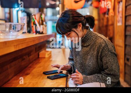 Signora asiatica in maglione casual utilizzando il telefono cellulare al banco nel tradizionale bar ramen Foto Stock