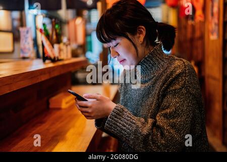 Vista laterale di una donna asiatica soddisfatta in un maglione casual sorridente mentre si utilizza il telefono cellulare al banco nel tradizionale bar ramen Foto Stock