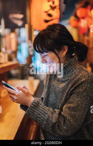 Donna asiatica in felpa casual sorridente mentre si utilizza il telefono cellulare al bancone nel tradizionale bar ramen Foto Stock