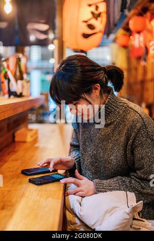 Signora asiatica in maglione casual utilizzando il telefono cellulare al banco nel tradizionale bar ramen Foto Stock