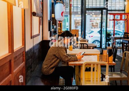 Signora asiatica in maglione casual utilizzando il telefono cellulare al banco nel tradizionale bar ramen Foto Stock