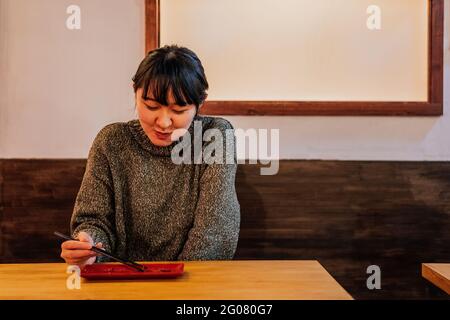 Giovane donna asiatica in maglione mangiare al bancone in legno nel caffè Foto Stock
