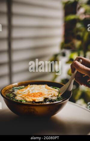 Mano di persona anonima che usa il cucchiaio per prendere yummy asiatico zuppa con tagliatelle e uova fritte sulla terrazza Foto Stock