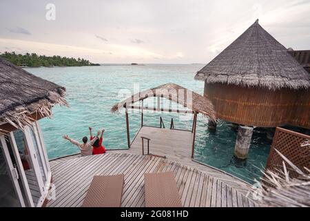 Dall'alto, vista posteriore di una donna irriconoscibile in abito rosso con braccio rialzato che balla vicino all'oceano con partner senza volto in abbigliamento casual rilassante alle Maldive Foto Stock