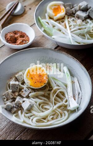 Tagliatelle di ramen fresche e cotte con tofu, uova e verdure servite su una ciotola di ceramica su un tavolo di legno Foto Stock