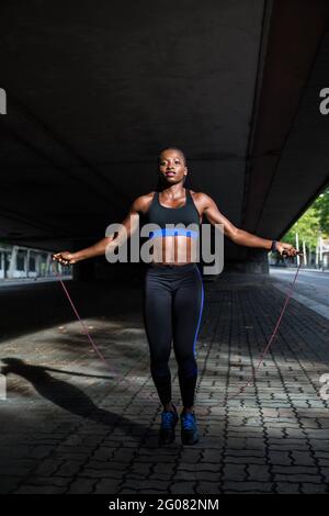 Donna afroamericana in abbigliamento sportivo con corda da salto e guardando la fotocamera mentre si è in piedi sul marciapiede sulla strada cittadina Foto Stock