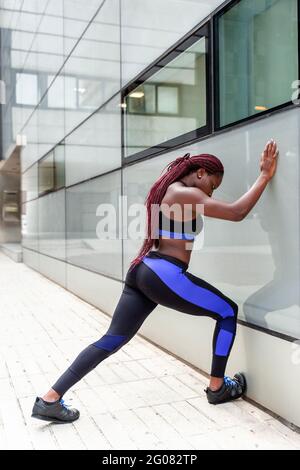 Vista laterale della giovane etnica muscolare Donna appoggiata al muro durante l'allenamento sulla strada Foto Stock