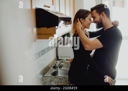 Allegro uomo bearded abbracciando sorridente Donna mentre si appoggia su armadio con lavandino in cucina accogliente a casa Foto Stock