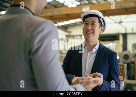 30s giovane uomo d'affari asiatico in tuta formale e elmetto mostrando partner commerciale straniero in background fabbrica. Partnership aziendale e concetto di successo del lavoro di squadra Foto Stock