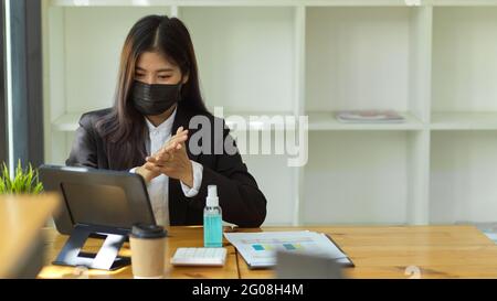 Donna d'affari con le mani della maschera medica igienizzante con gel di alcool prima di iniziare a fare il suo lavoro Foto Stock