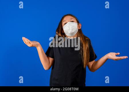 Bella ragazza pre-adolescente con una maschera chirurgica per proteggersi dal coronavirus che le pone le mani in su in un gesto di dubbio. Concetto di gesto Foto Stock