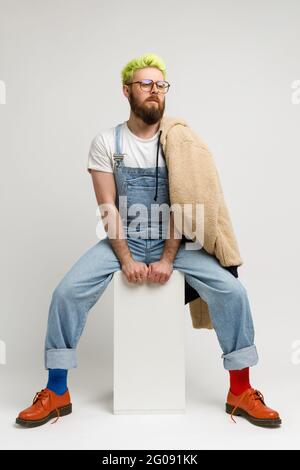 Foto a tutta lunghezza di un uomo modello elegante che indossa abiti alla moda e scarpe rosse, seduto su un cubo bianco, guardando lontano con un'espressione sognante. studi interni Foto Stock