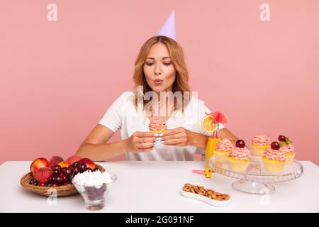 Ritratto di bella giovane donna adulta che tiene torta di compleanno e soffia candela, celebrazione da sola, facendo desiderio, femmina in T-shirt casual e cono di festa Foto Stock