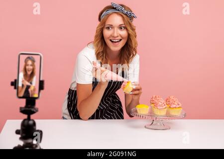 Ritratto del blogger di cibo felice eccitato che trasmette in diretta, mostrando alla telecamera del processo di decorazione delle torte, utilizzando cono di pasticceria. Indù Foto Stock