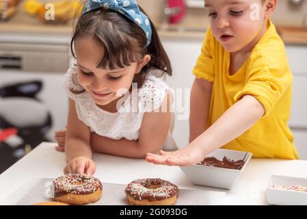 Bambini che decorano ciambelle con spruzzi colorati in cucina a casa Foto Stock