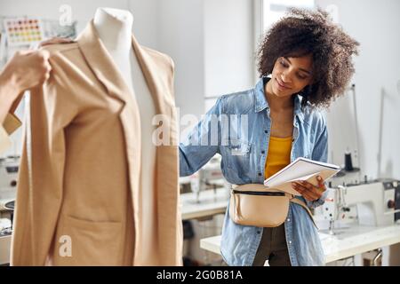 Donna afroamericana con notebook guarda l'elegante giacca beige mentre il designer misura i dettagli in studio Foto Stock