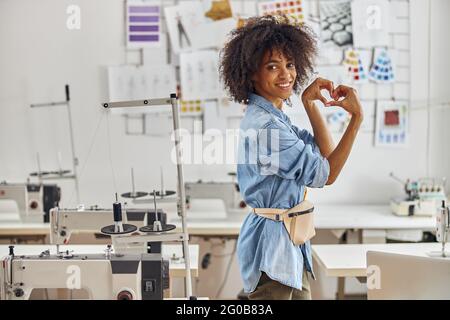 Afroamericano designato mostra cuore con le mani vicino macchina da cucire in officina Foto Stock