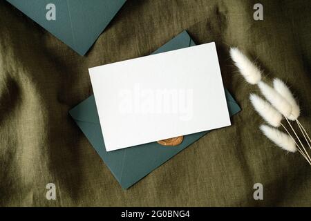 Set di cancelleria per matrimoni. Carta d'invito bianca mockup e fiori secchi su tessuto verde vintage. Disposizione piatta, vista dall'alto, spazio per la copia. Foto Stock