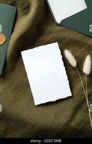 Carta bianca di invito mockup con buste e fiori secchi su tessuto verde. Set di cancelleria vintage per matrimoni. Disposizione piatta, vista dall'alto, spazio per la copia Foto Stock