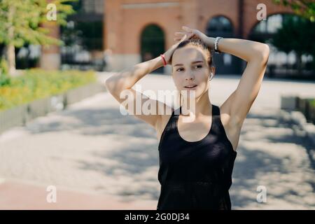 Donna sportiva concentrata in distanza respira profondamente dopo che il jogging prende la pausa durante l'allenamento cardio vestito in t-shirt nera tiene le mani sulla testa Foto Stock