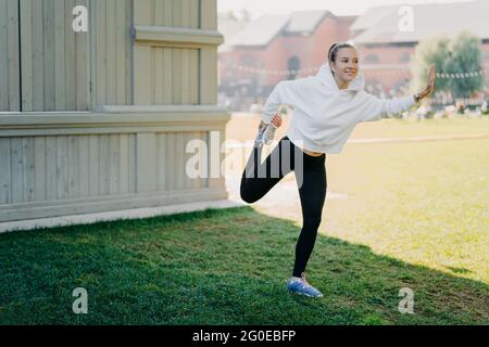 Felice giovane donna attiva in felpa con cappuccio e leggings si prepara per running stratches e scalda le gambe focalizzate felicemente a distanza ascolta la musica via Foto Stock