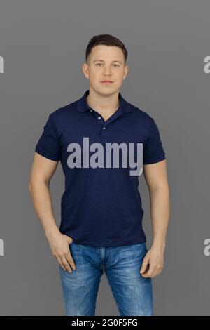 Il giovane serio si erge su sfondo grigio in polo e jeans. Concetto di auto-espressione e raggiungimento di obiettivi. Foto Stock