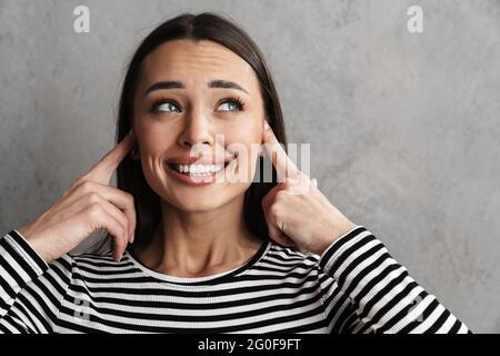 Primo piano di una giovane donna sorridente attraente che blocca le orecchie isolate su sfondo grigio Foto Stock