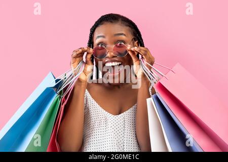 Signora nera fredda con grappoli afro che tengono le borse della spesa, occhiali da sole toccanti, non può credere grande vendita, sfondo rosa Foto Stock