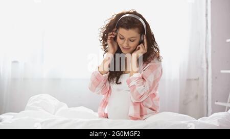 donna incinta sorridente in cuffia che ascolta musica in camera da letto Foto Stock