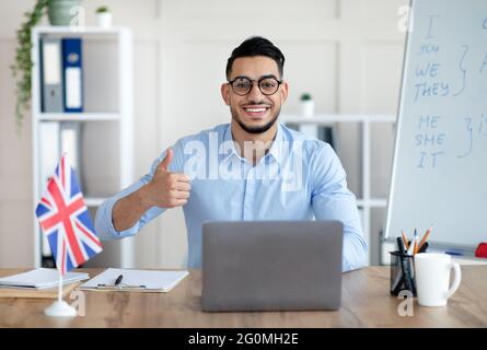 Giovane tutor arabo che conduce una lezione di inglese online, mostrando il pollice in su, insegnando la lingua straniera da casa Foto Stock
