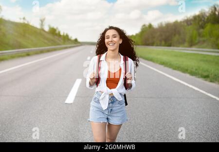 Gioiosa giovane donna in abiti estivi casual a piedi lungo strada asfaltata in campagna, hitchhiking per passeggiate all'aperto Foto Stock